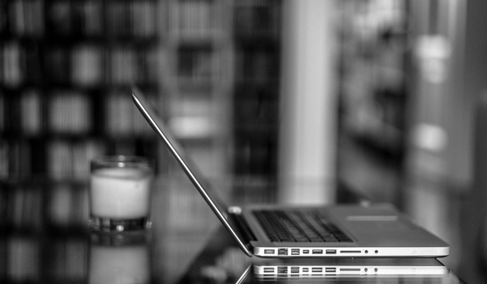 Black and white image of a laptop on a reflective glass table in a blurred room setting.